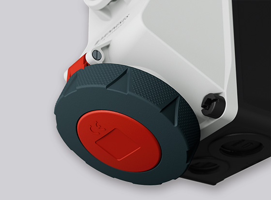 Die weiße Wandsteckdose DUO besitzt ein schwarzes Gehäuseunterteil und eine nach unten gerichteten Anschluss, wobei der runde Klappdeckel außen grau und in der Mitte rot ist.